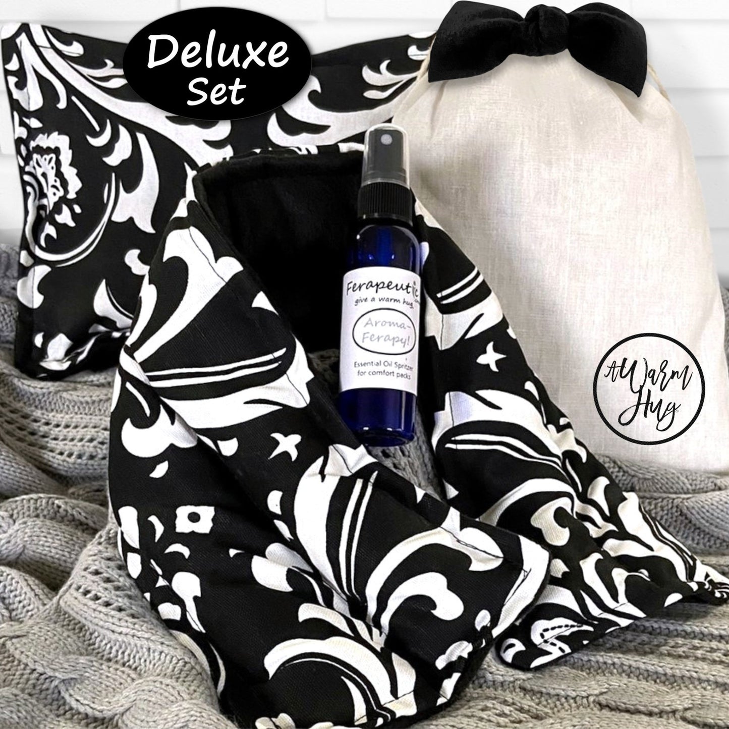 Deluxe set with black and white patterned heat packs, bottle of aromatherapy, and white gift bag with black bow on a textured surface.