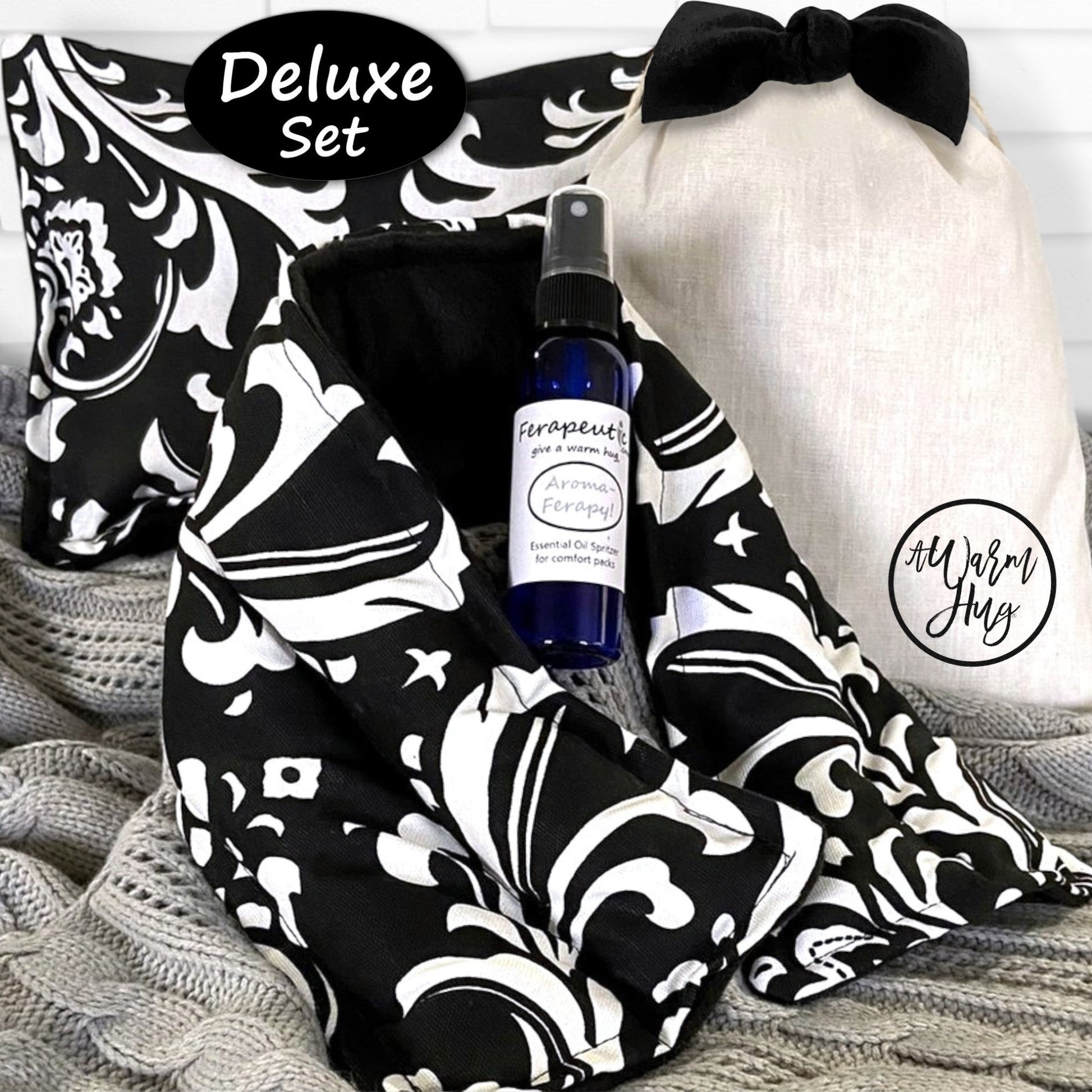 Deluxe set with black and white patterned heat packs, bottle of aromatherapy, and white gift bag with black bow on a textured surface.