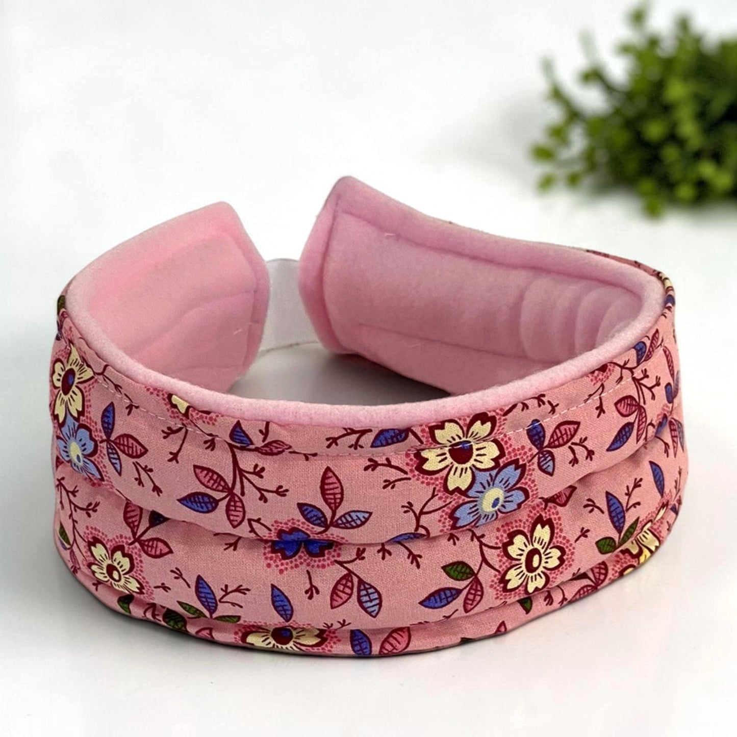 A padded pink headband with a floral print to be used hot or cold for headaches, tmj and jaw pain.  It is featuring a soft pink velvet-like inner lining and a fabric outer band decorated with yellow, blue, and purple flowers on a pink background.