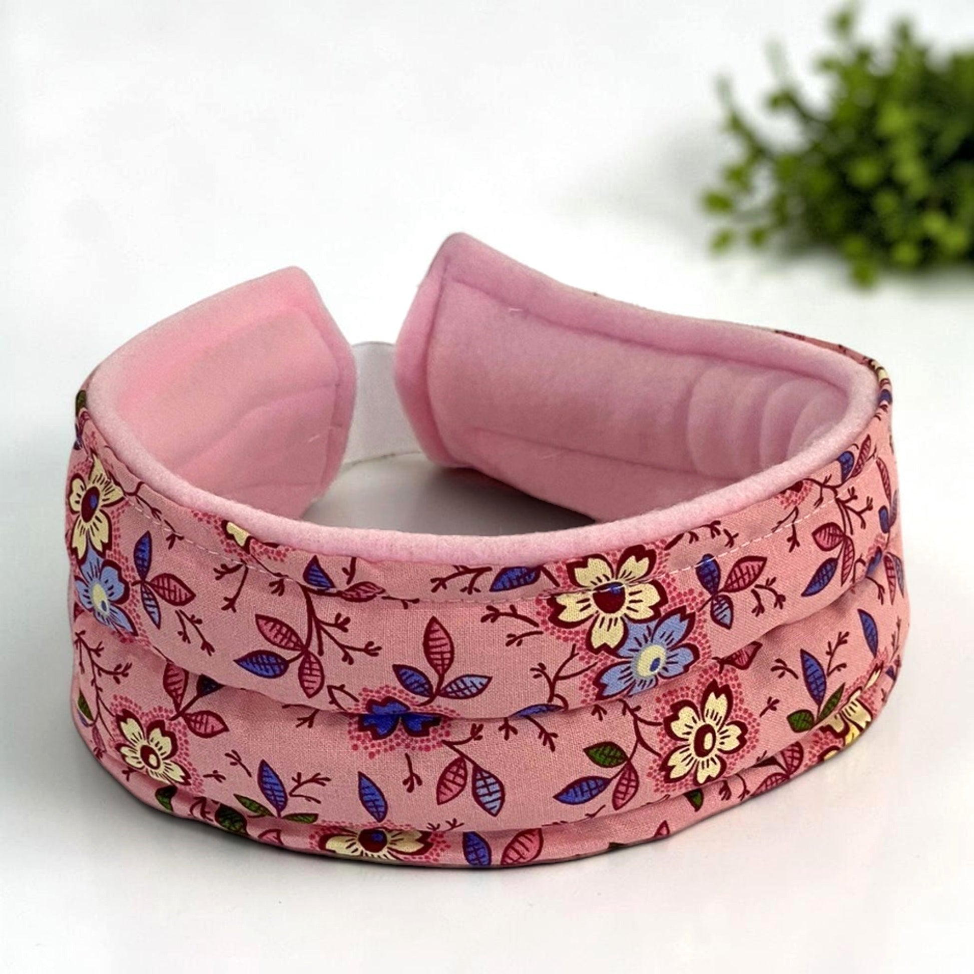 A padded pink headband with a floral print to be used hot or cold for headaches, tmj and jaw pain.  It is featuring a soft pink velvet-like inner lining and a fabric outer band decorated with yellow, blue, and purple flowers on a pink background.