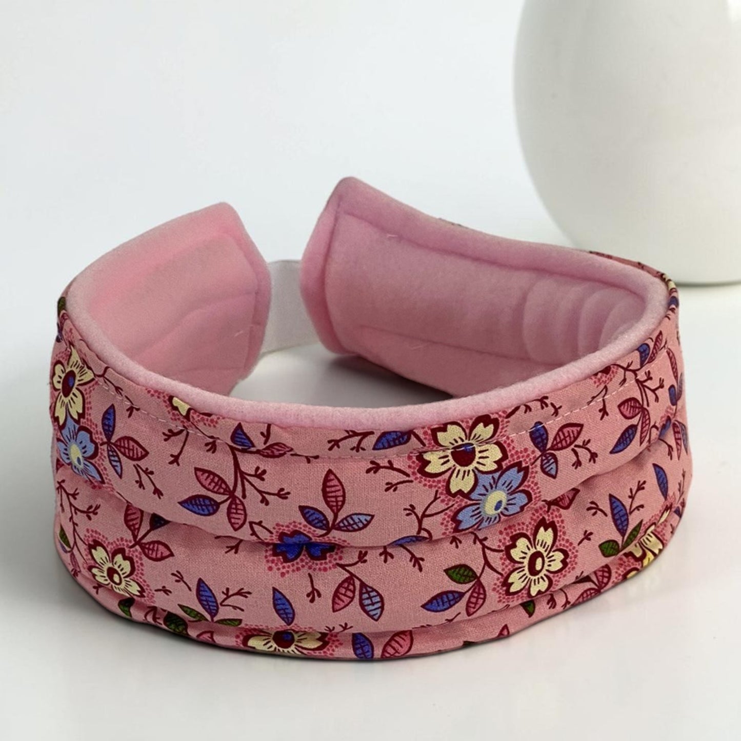 A heating pad pink headband with a floral print on the exterior and a solid pink interior, displayed on a white surface.