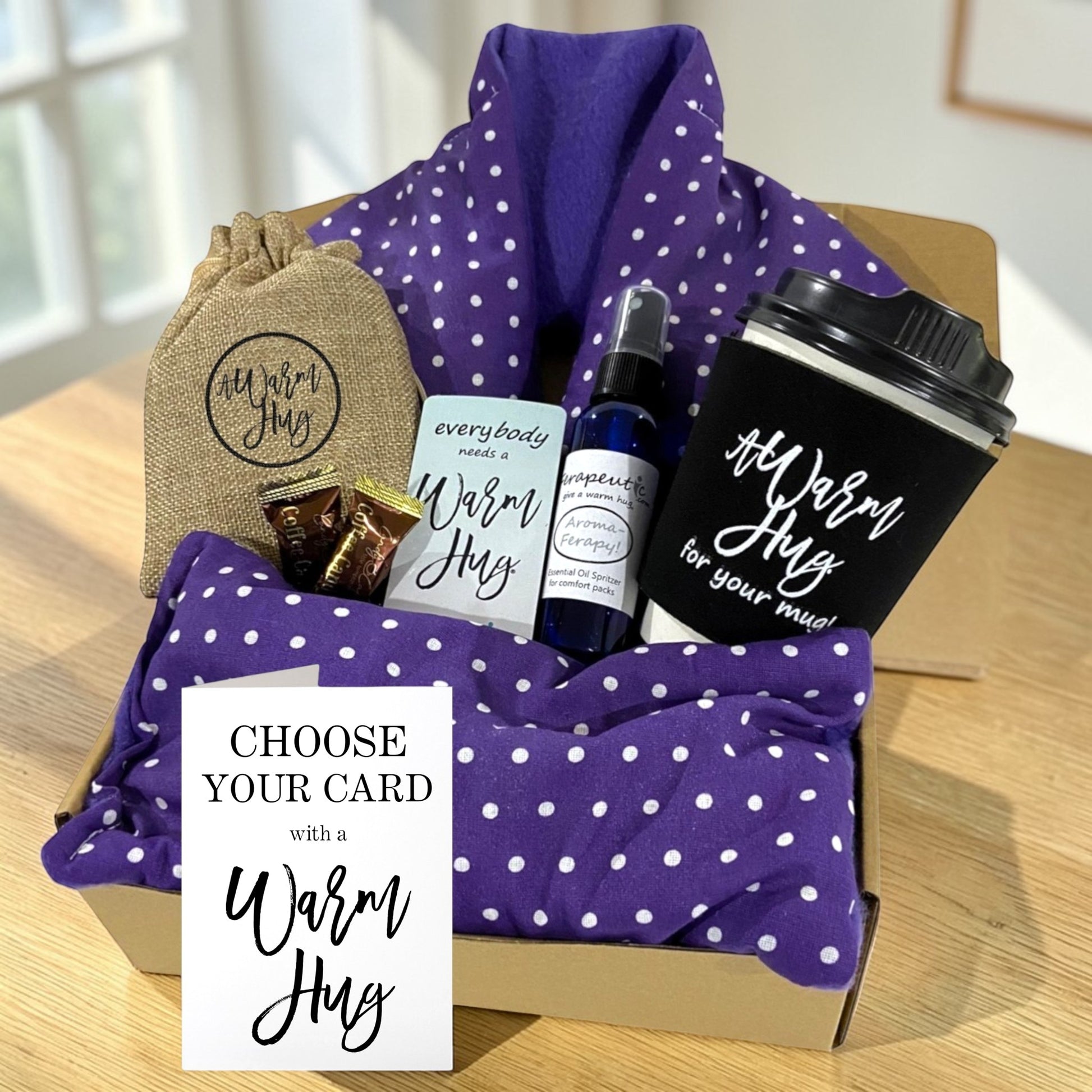 Gift set with purple polka dot microwave heating pad, coffee cup, and other items with a warm hug gift card on a wooden surface.